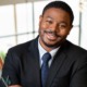 Ali Knight newsmaker headshot: black man in suit smiling while leaning to his left in front of bright background
