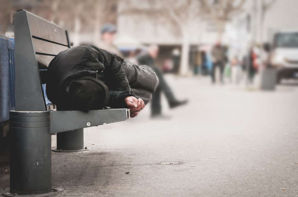 NY Homeless Payments: Person in dark clothes sits cross-legged on city bench huddled over