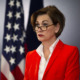 Child Labor Laws: Iowa Governor Kim Reynolds, a woman with short dark hair and dark-framed glasses, wearing red suit with white blouse, standing next to American flag while speaking into microphone.