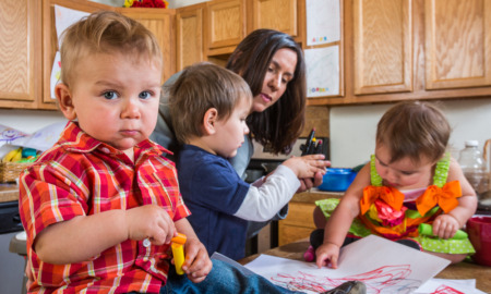 Childcare Utah: Woman caring for three toddlers sitting on kitchen island counter doing an art project in kitchen and baby in red plaid shirt with guilty face in foreground looks at camera