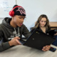 HS Data Science vs Math: Two older teens, both in dark long-sleeved swearshirts, sit at school desk, one Black bot with laptop in hand and one girl looking at laptop screen