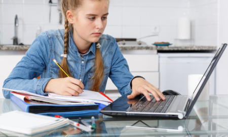 Oklahoma virtual charter schools: Teenage girl with ling blonde hair in two braids in light blue denim shirt sits in white kitchen at table with several school supplies working on laptop and writing in notebook