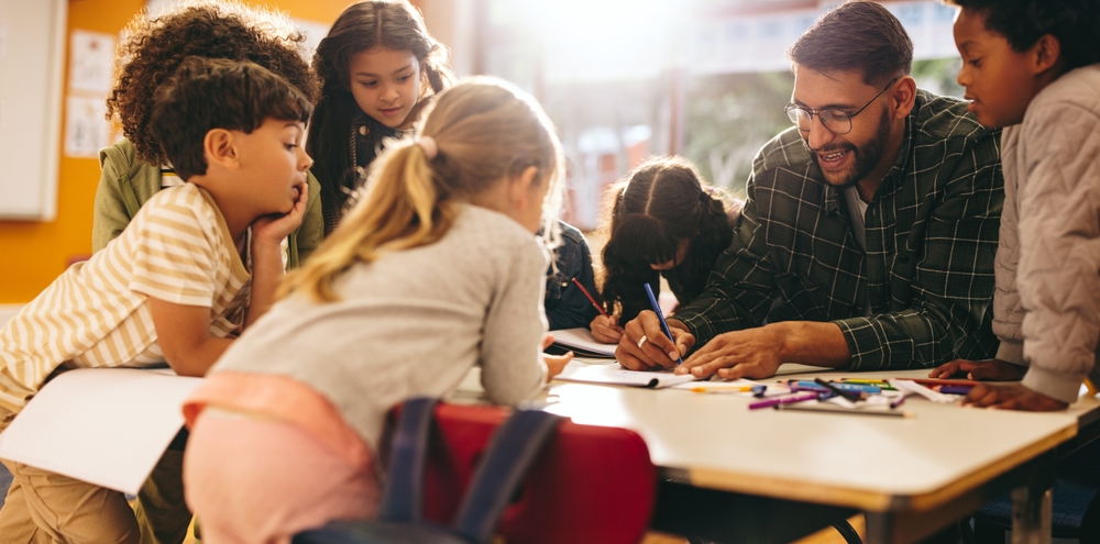 Teacher layoffs: Group of elementary school kids stand around a table in a classroom, attentively leaning over table and following a lesson being taught by their male teacher with glasses, dark hair and beard.