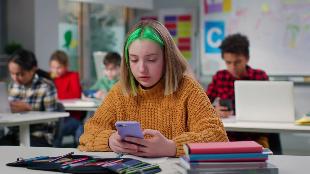 School cell phone bans: Teenage schoolgirl with green hair chatting on smartphone sitting at desk in class. Teen student using cellphone in classroom at school