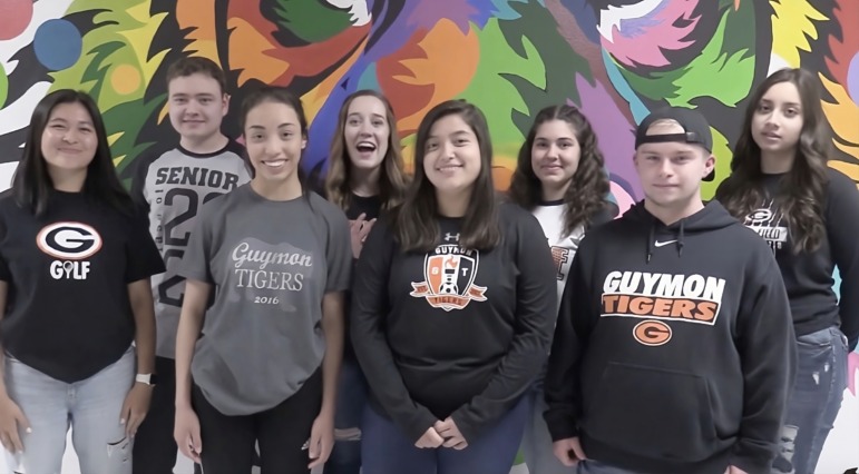 Immigrant student school admissions: 8 teen students - all wearing black or white tops with touches of orange - stand in 2 rows in front of a very colorful mural of abstract tropical plants