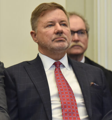 Oklahoma-strikes-down-religious-charter-school: Several people sit in rows of chairs. White man with blonde hair in in center photo wearing gray suit, white shirt and red tie with hands in pants pockets.