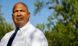 Uvalde indictment: Bald white man in white business shirt and dark tie stands staring seriously into camera under blue sky with green trees in the background.