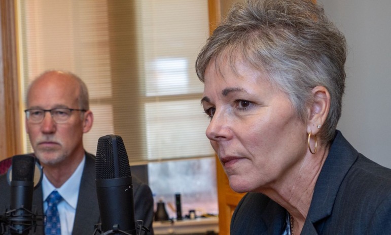 Kansas literacy teacher training: Two older adults in dark business suits — one woman, one man — sit at table speaking into microphones.