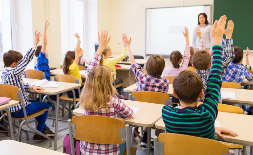 Segregation academies: Back view of group of all white school kids sitting at classroom desks and raising hands with white female teacher standing facing students
