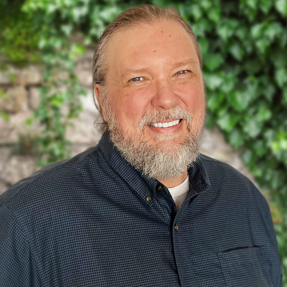 Lie detector tests and juveniles: Headshot young, smiling, middle-aged man with combed back graying blonde hair in navy shirt standing in front of green foliage on wall