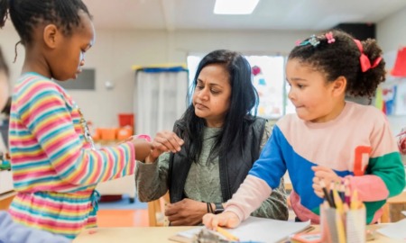 Universal prekindergarten is coming to California: woman with long, dark hair helps two young girls in preschool classroom