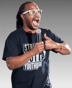 Jamiel Alexander Headshot: Black manwith long hair and clear-framed glasses in black t-shirtstands speaking with hands acoss chest as fists meetingt